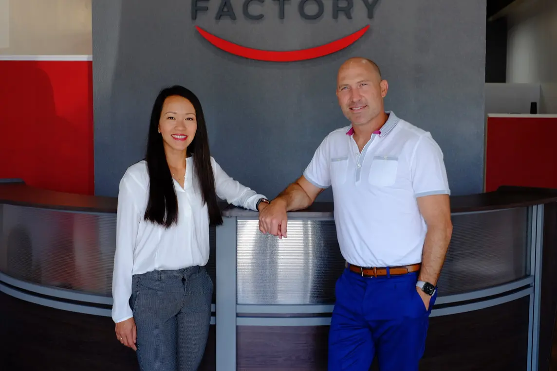 Dr. Trang Cao and Dr. Marty Greenberg, orthodontist experts at Bold Bite Orthodontics in Jacksonville FL, smile in their clinic. Dr. Cao wears a white blouse, Dr. Greenberg a white polo. They stand by a reception desk.