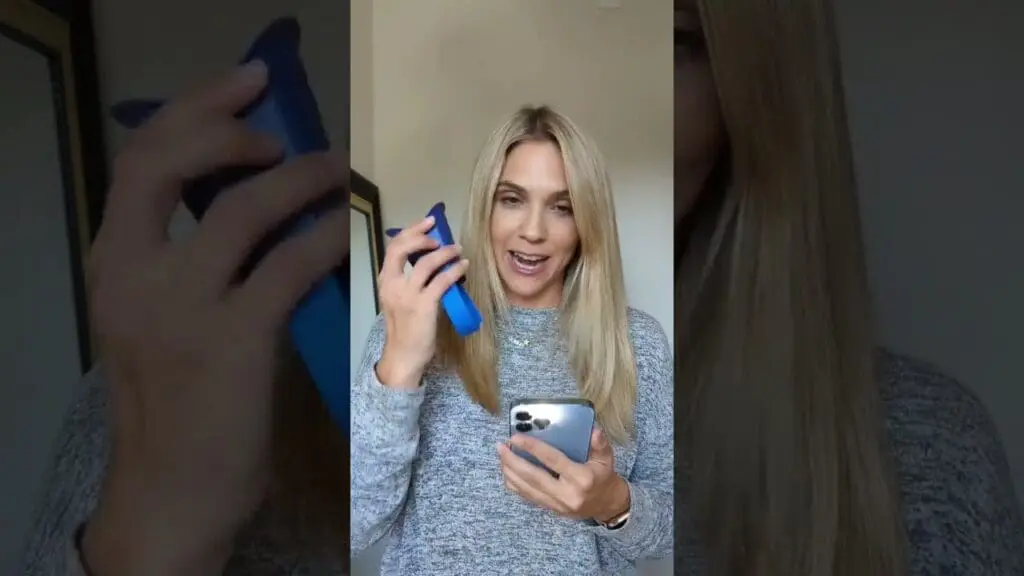 A person with long blonde hair holds a blue phone case and a silver smartphone, looking at the camera against a neutral background.