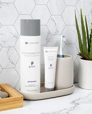 Toothbrush and toothpaste in a ceramic holder next to a bottle of maintenance rinse and a small potted plant on a bathroom counter with hexagonal tile backsplash.