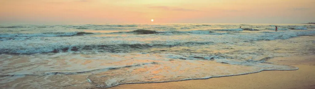A beach scene with gentle waves washing onto the shore at sunset. The sky is a mix of soft pinks and oranges, and a person is visible in the distance near the water. A beach scene with gentle waves washing onto the shore at sunset. The sky is a mix of soft pinks and oranges, and a person is visible in the distance near the water.