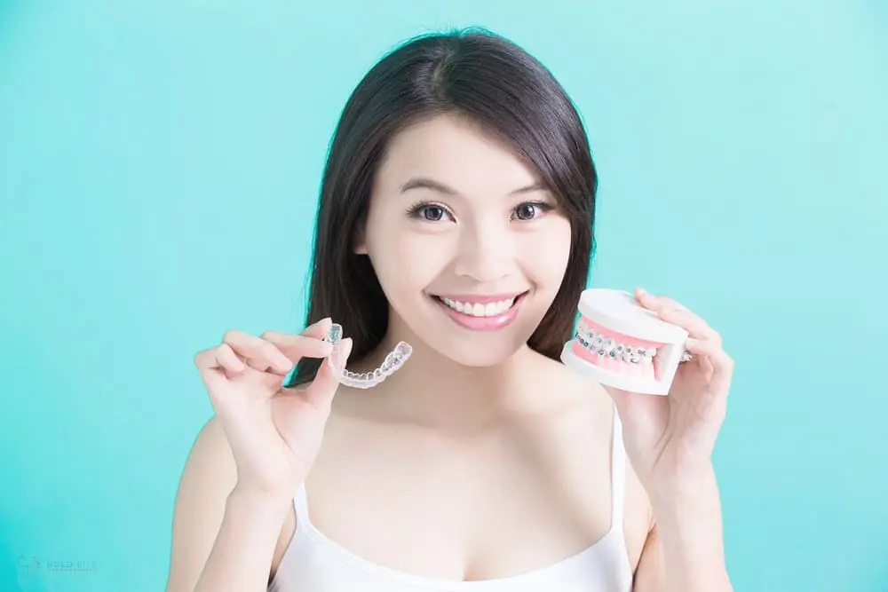 Person holding a dental aligner in one hand and a dental model with braces in the other, smiling against a turquoise background, contemplating Invisalign cost in Jacksonville, FL options.