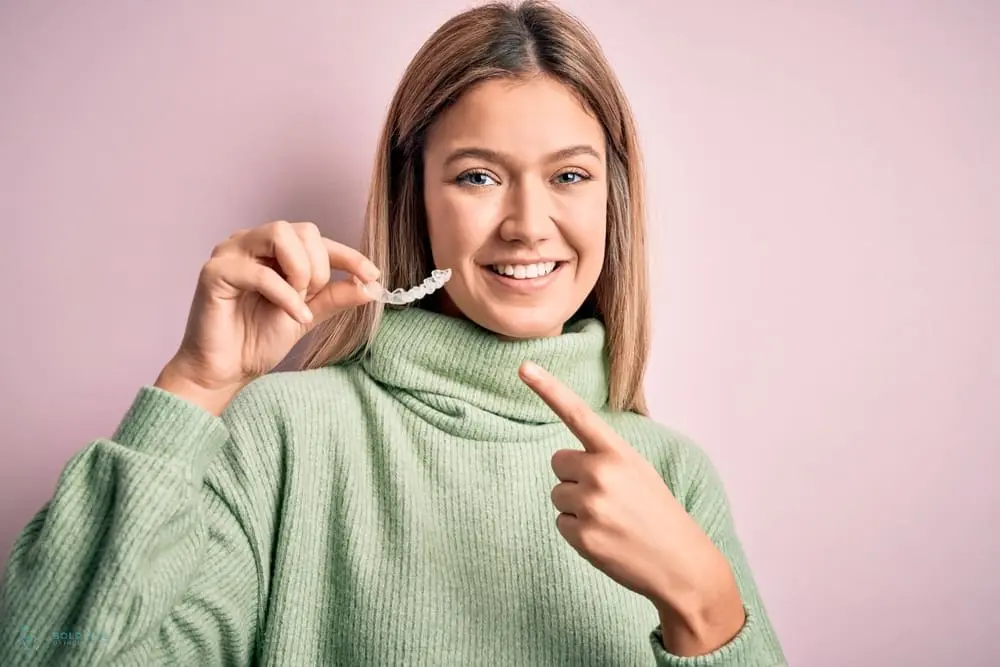 A person wearing a green sweater holds up a clear dental aligner and points to it, smiling against a pink background, perhaps considering the Invisalign cost in Jacksonville, FL.