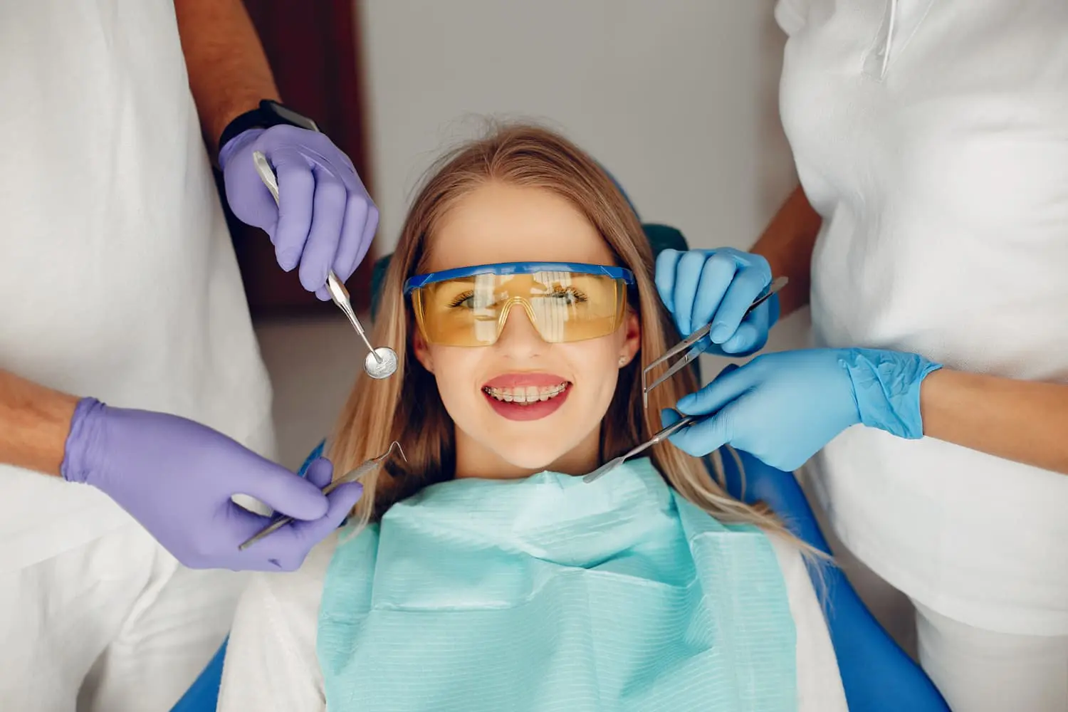 A woman wearing clear braces protective glasses smiles in a dental chair. Two people wearing gloves hold dental tools near her mouth.