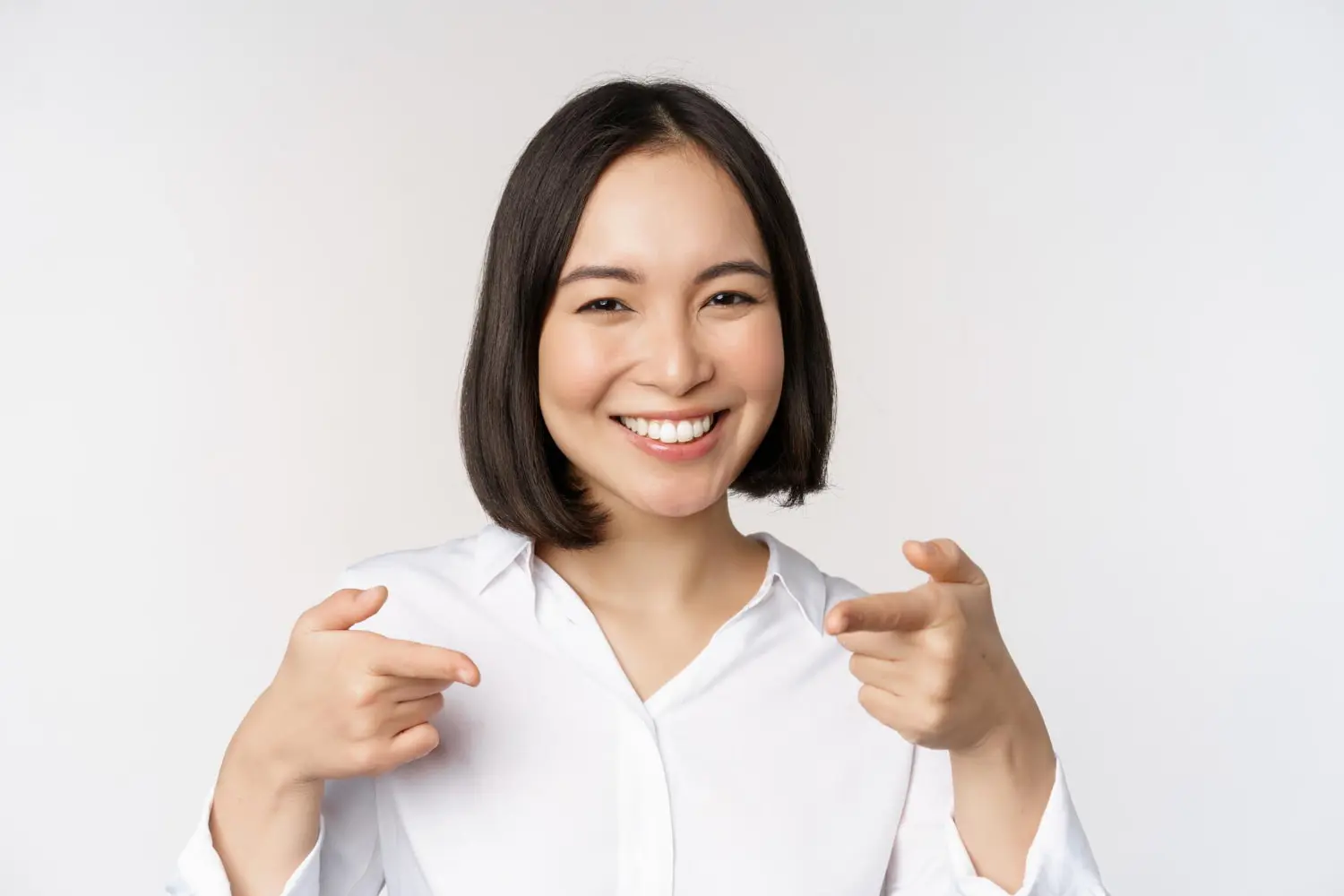 Smiling adult person with short black hair and a white shirt points both index fingers forward against a plain background at Bold Bite orthodontics.