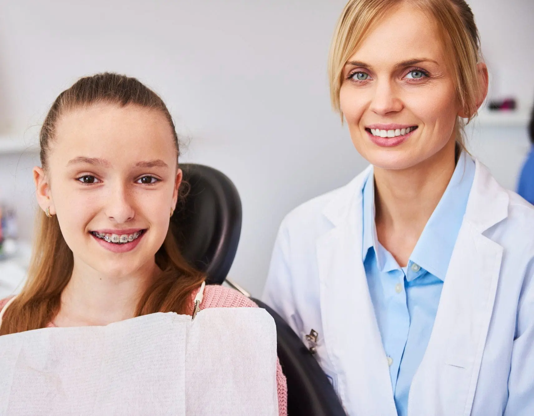 A girl with metal braces sits in a dentist’s chair next to a smiling dentist in a white coat in Jacksonville, FL.
