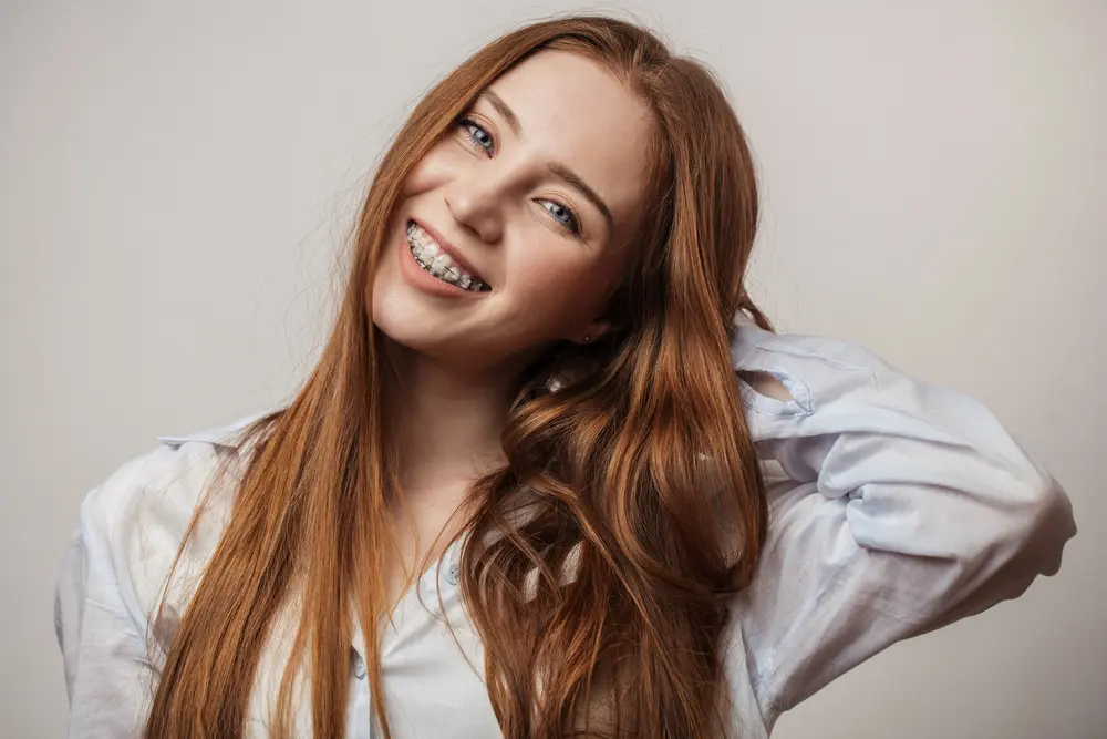 Bold Bite Orthodontics A smiling woman with long red hair and braces, wearing a light blue shirt, poses against a plain background, possibly showcasing clear braces in Jacksonville, FL.