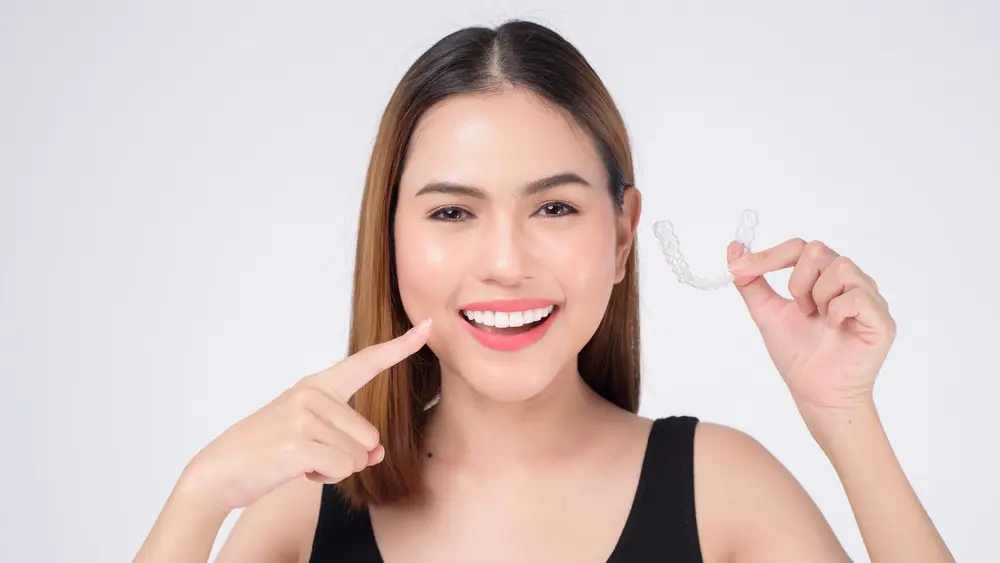 Person smiling while holding Invisalign clear dental aligner and pointing to their teeth, against a plain white background in Jacksonville FL