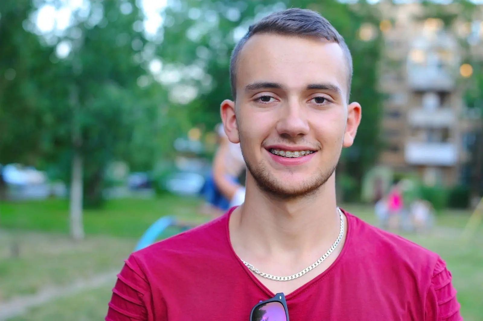 A young man with clear braces and short hair smiles at the camera, wearing a red shirt and a silver necklace. Trees and a building are blurred in the background in Jacksonville, FL.