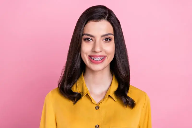 A person with long dark hair and metal braces smiling, wearing a yellow shirt against a pink background in Jacksonville, FL.