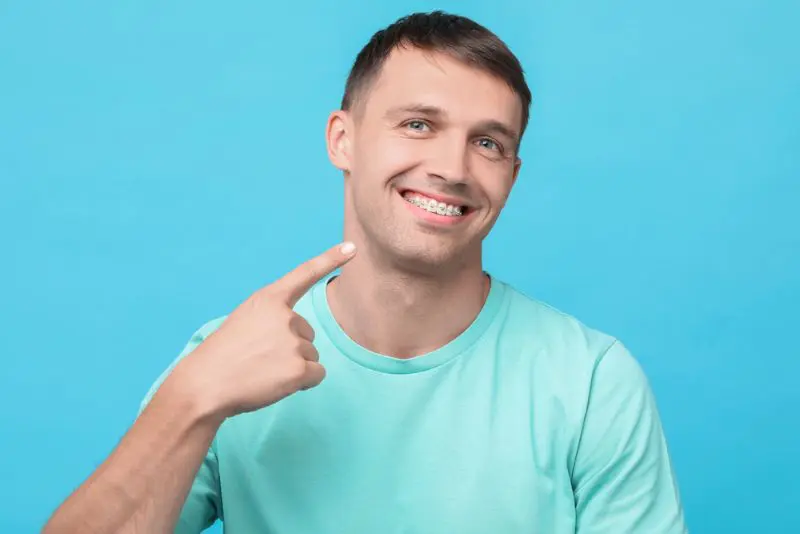 Man in a teal shirt smiles and points at his metal braces against a solid blue background in Jacksonville, FL.