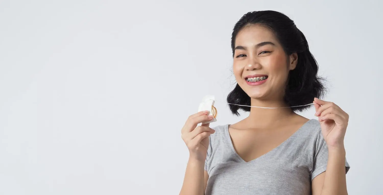 A person with metal braces smiling, holding dental floss in both hands against a plain background in Jacksonville, FL.