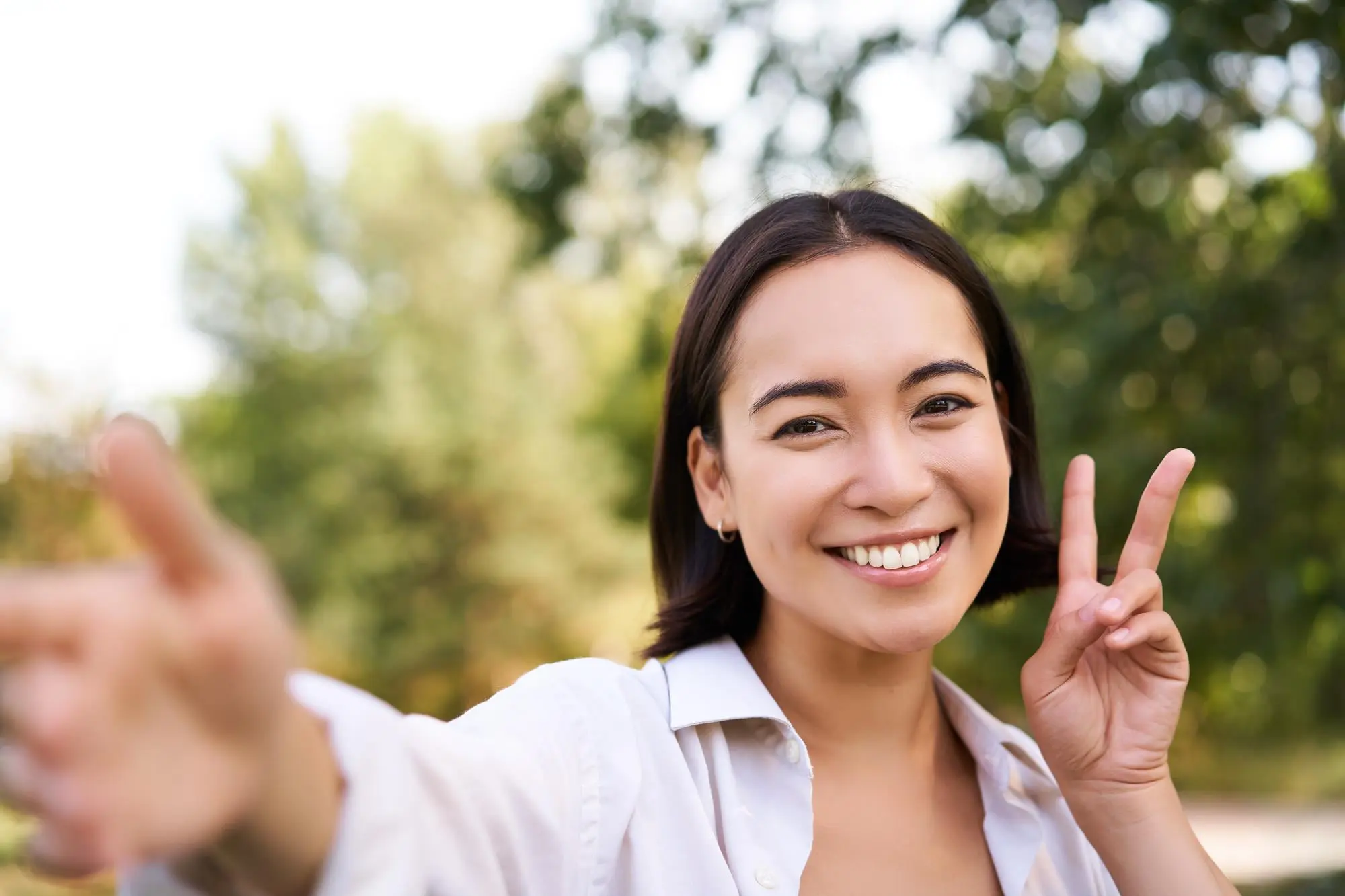 A woman outdoors in FL smiles at the camera, making a peace sign with one hand and pointing with the other. Green trees are visible in the blurred background, capturing her excitement about finding affordable braces near me in Jacksonville.
