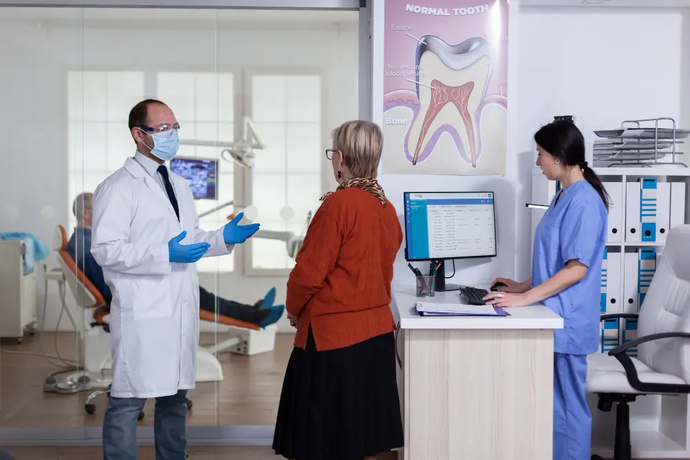A dentist wearing a mask and gloves talks to a woman in an orange sweater about orthodontic insurance plans while a nurse works at a computer in a dental clinic. A patient sits in a chair in the background in Jacksonville, FL.