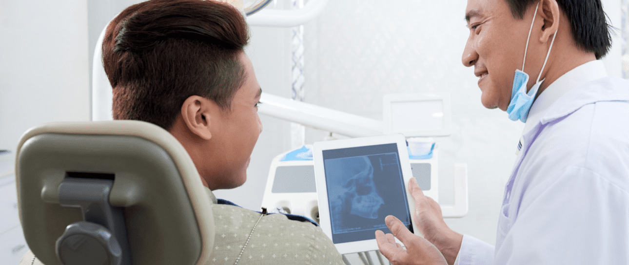 A dentist shows a dental X-ray on a tablet to a seated patient in a dental clinic, possibly discussing the results of jaw surgery in Jacksonville, FL.