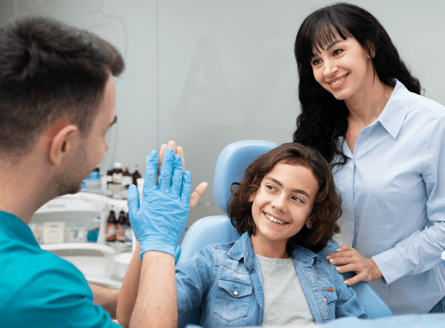 A dentist wearing gloves gives a high five to a smiling child sitting in a dental chair, with an adult woman standing nearby, likely celebrating successful recovery from jaw surgery in Jacksonville, FL.