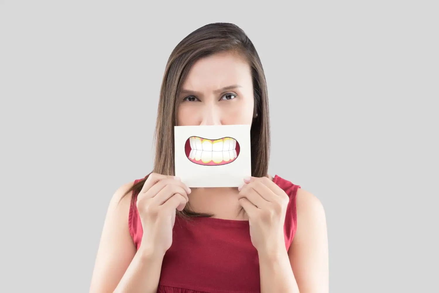 In Jacksonville FL, a woman in a red sleeveless top holds a card with an illustrated wide smile in front of her mouth, playfully highlighting her underbite against a plain gray background.