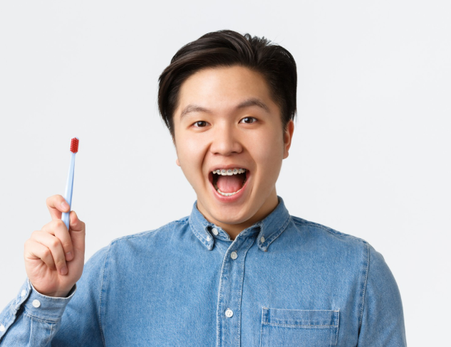 Young person in a denim shirt holding a toothbrush and smiling with mouth open, showing adult braces, against a plain background in Jacksonville, FL.