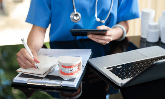 A person in scrubs writes notes on orthodontic insurance plans in a notebook with a dental model nearby, while holding a phone next to a laptop on a desk in Jacksonville, FL.