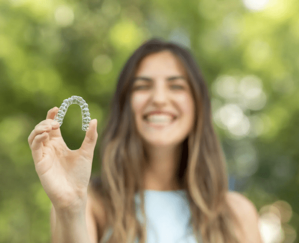 A woman holds a clear dental aligner and smiles, possibly discussing Invisalign insurance options, with blurred green foliage in the background in Jacksonville, FL.