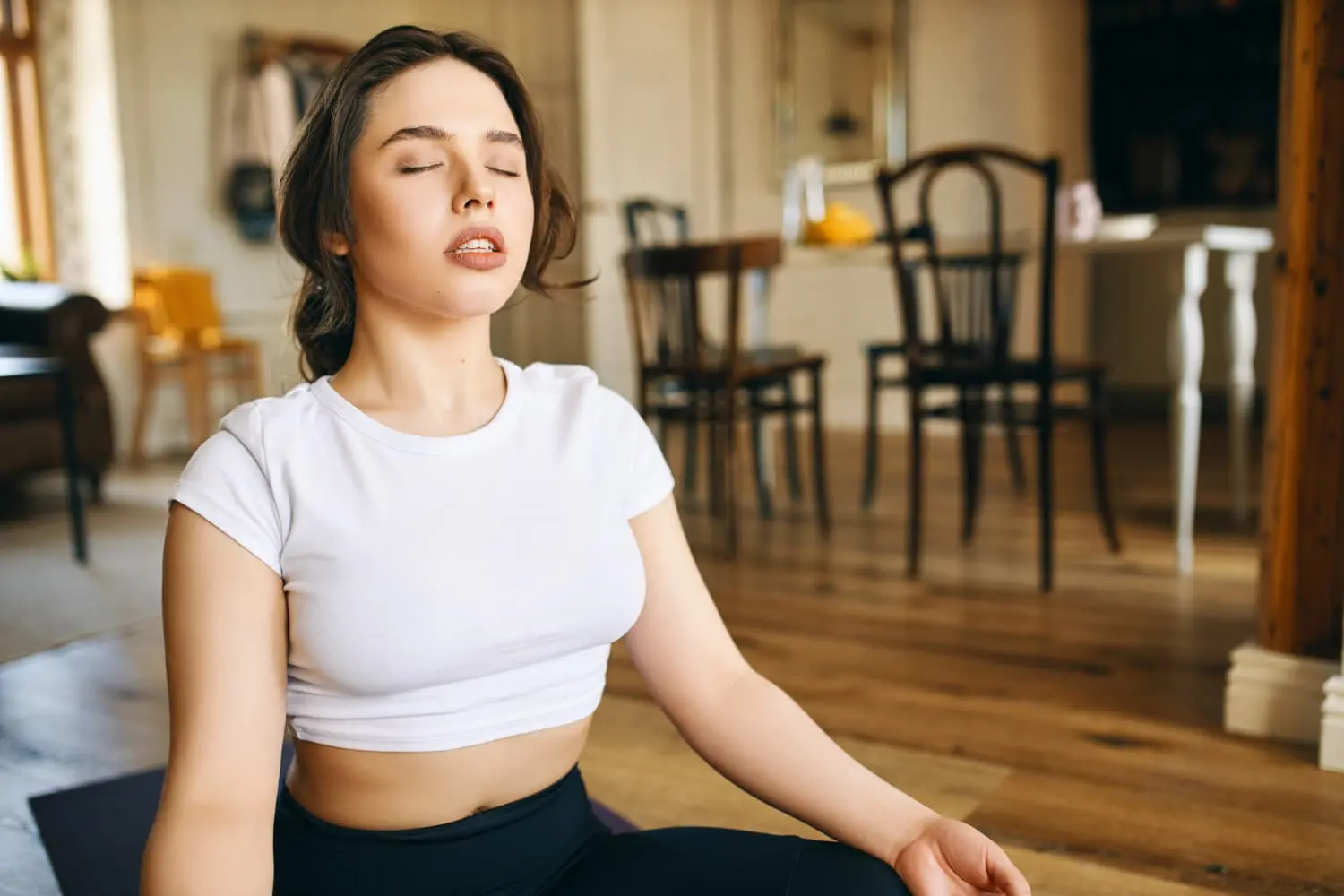 In Jacksonville FL, a woman in a white crop top sits cross-legged indoors with eyes closed, practicing deep breathing techniques often recommended to help mouth breathers transition to nasal breathing.
