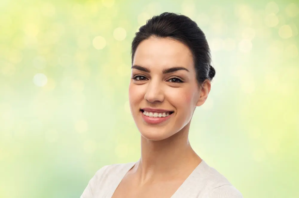 A woman with dark hair pulled back smiles at the camera, standing in front of a green, blurred FL background with light spots—just like the fresh confidence after getting Clear Braces in Jacksonville.