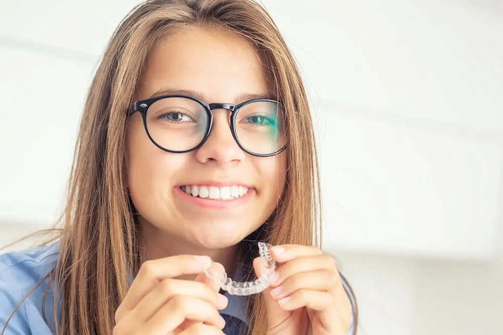 A young person with long hair and glasses is smiling while holding a clear dental retainer in both hands in Jacksonville, FL. If you're interested in maintaining your smile, ask us about our retainer options and how we can help you keep your teeth aligned in Jacksonville!