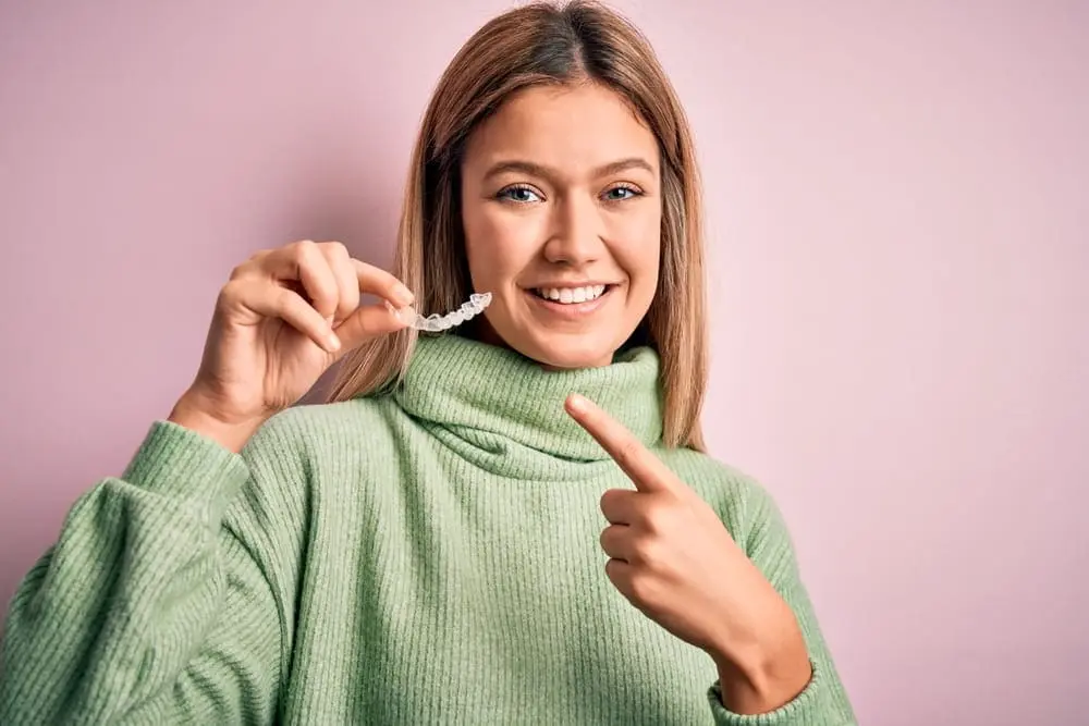 A young woman in a green sweater smiles and points at a clear dental aligner she is holding against a light pink background in Jacksonville, FL. If you're interested in improving your smile, ask about our Invisalign options and flexible payment plans available in Jacksonville!