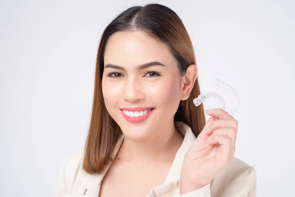A woman with straight brown hair smiles while holding a clear dental aligner in her right hand, possibly discussing Invisalign insurance coverage in Jacksonville, FL.
