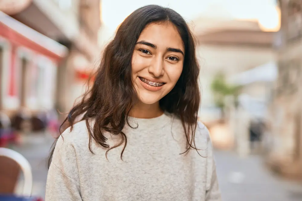 Young woman with long brown hair and metal braces in Jacksonville, FL, smiles while standing outdoors on a city street; she is wearing a light grey sweater.