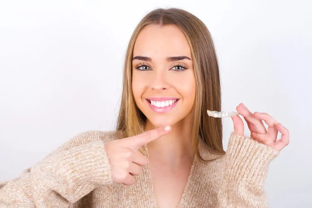 A young woman with long hair smiles and points to a dental aligner she is holding in her hand, highlighting the benefits of an Invisalign Payment Plan in Jacksonville FL, while standing against a plain white background.
