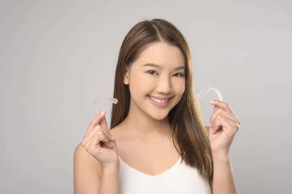 A woman smiles while holding two clear dental aligners in each hand, highlighting the benefits of an Invisalign Payment Plan in Jacksonville FL against a plain background.