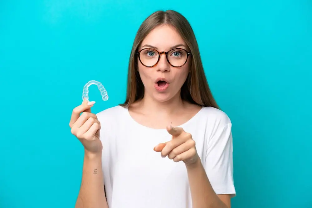 A young woman with glasses holds a clear dental aligner in one hand and points forward with the other, possibly exploring Invisalign insurance options for treatment in Jacksonville, FL.