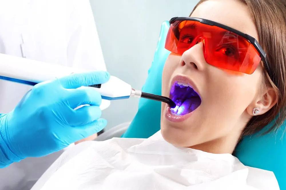 A dentist uses a curing light tool inside a patient's mouth, who is wearing protective red glasses and a dental bib in Jacksonville, FL, highlighting the treatment process and considerations between Invisalign vs braces for orthodontic care.