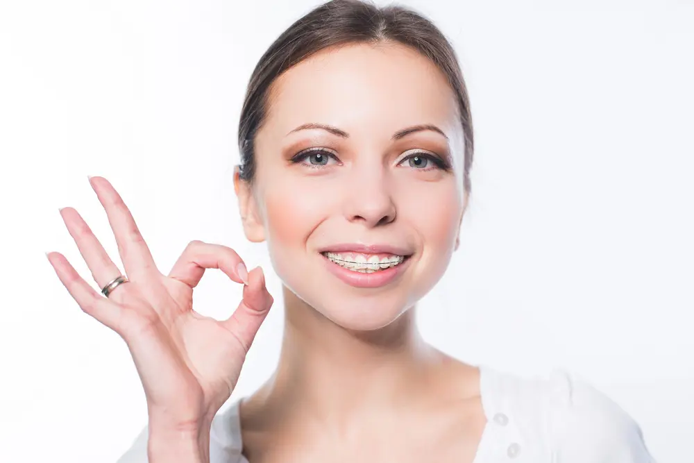 A woman with braces smiles and makes an "OK" hand gesture. She is wearing a white top and a ring on her finger. The background is white, highlighting confidence after addressing causes of severe overjet and overbite in Jacksonville FL.