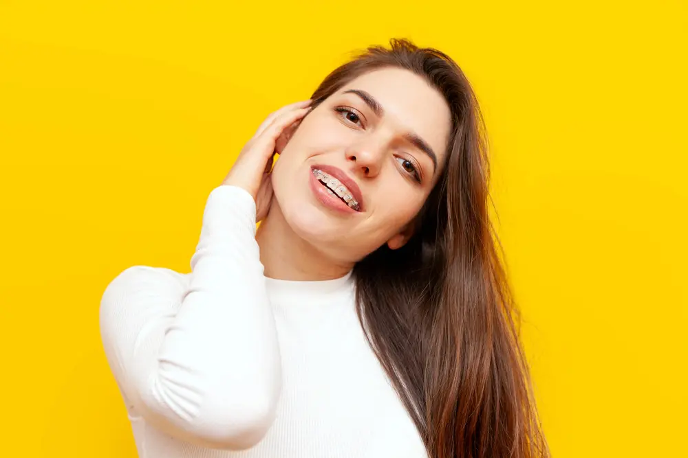 Young woman with long brown hair, wearing a white shirt and our clear ceramic braces, smiling and touching her neck, stands against a bright yellow background in Jacksonville, FL.