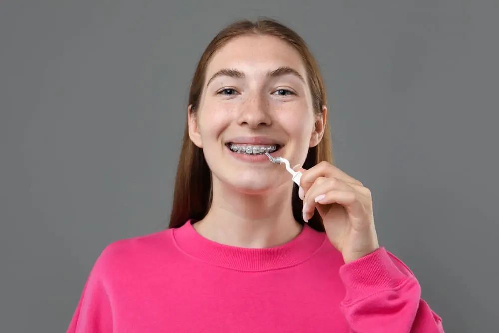 A young person with braces uses a dental floss pick to clean their teeth, standing against a plain gray background and wearing a pink sweatshirt in Jacksonville FL, emphasizing the importance of oral hygiene while considering options like Invisalign vs braces.