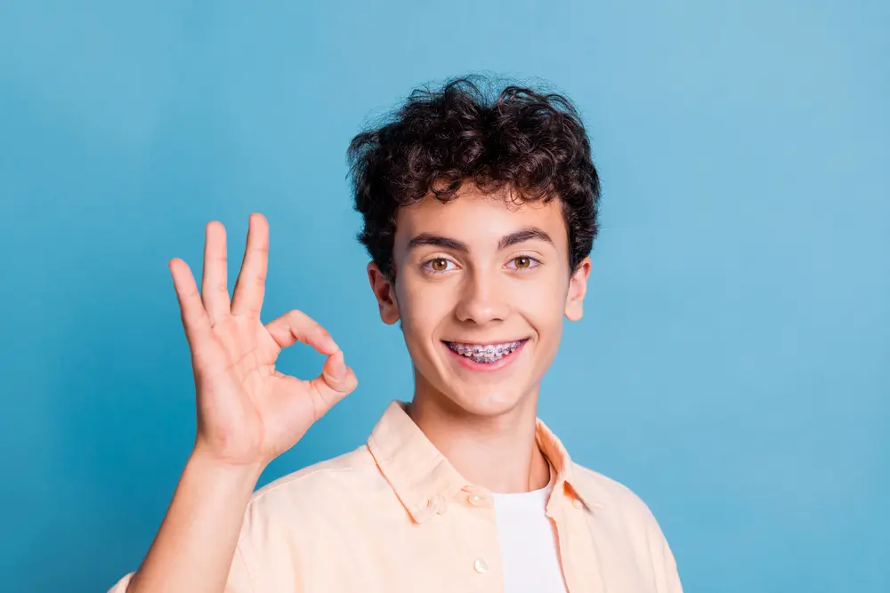 Teenager with curly hair and braces showing an OK hand gesture, smiling confidently, and standing in front of a plain blue background—perfectly capturing the journey of correcting an overbite in Jacksonville FL.