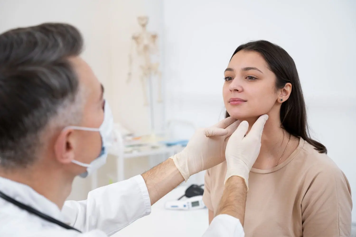 A doctor wearing gloves and a mask examines a young woman’s neck in a medical office, possibly assessing her recovery after jaw surgery in Jacksonville, FL.