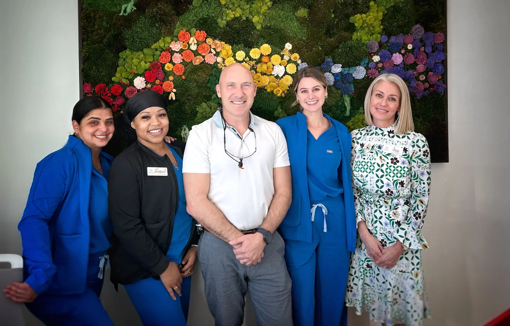 Five people, including three in blue scrubs, one in a black jacket, and one in a white dress, stand smiling in front of colorful floral wall art as the Bold Bite Orthodontics team in Jacksonville, FL.
