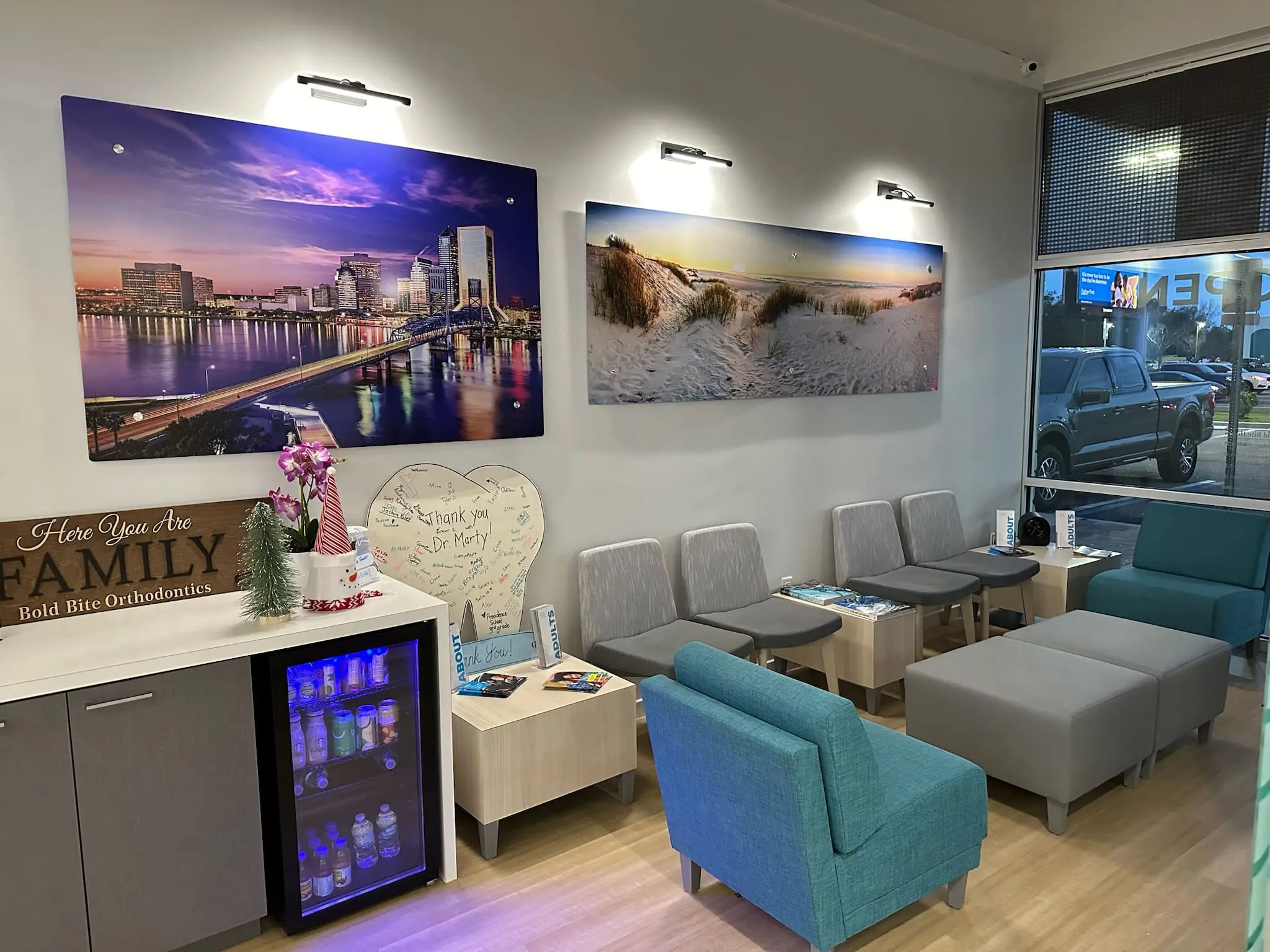 A modern and comfortable waiting room with gray and teal seating, a refreshment station, wall art, and magazines on tables at Bold Bite Orthodontics in Jacksonville, FL.