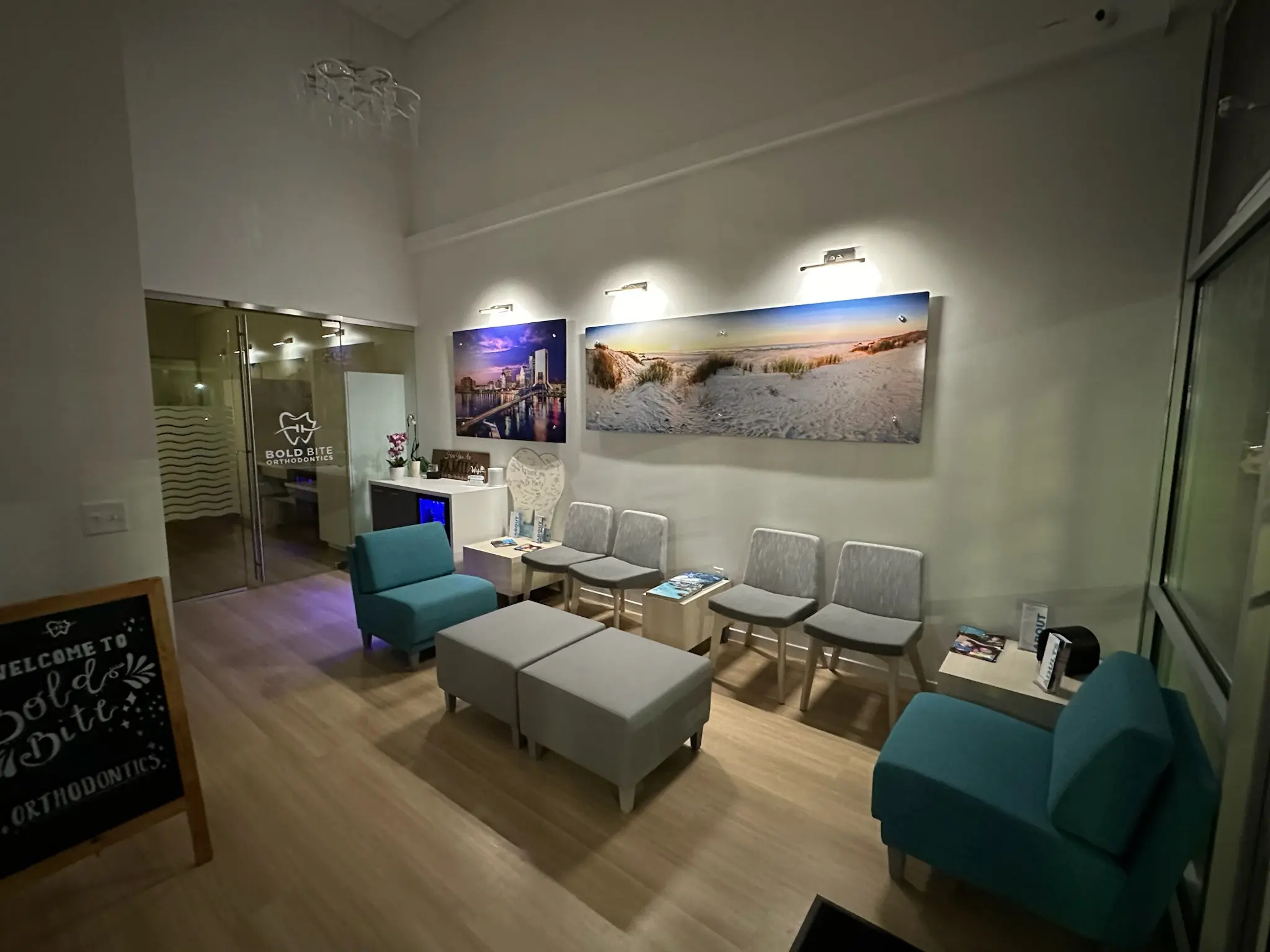 A modern waiting room at Bold Bite Orthodontics in Jacksonville, FL features gray and teal chairs, beach-themed wall art, a chalkboard sign, magazines on a table, and a glass door labeled "Bold Bite Orthodontics.