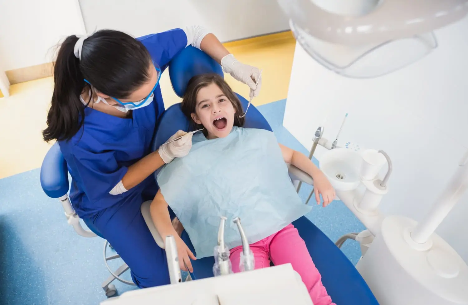 A dental professional examines a young girl’s open mouth with orthodontic appliances visible in a dental chair; the girl wears a bib, and dental tools are visible nearby in Jacksonville, FL.