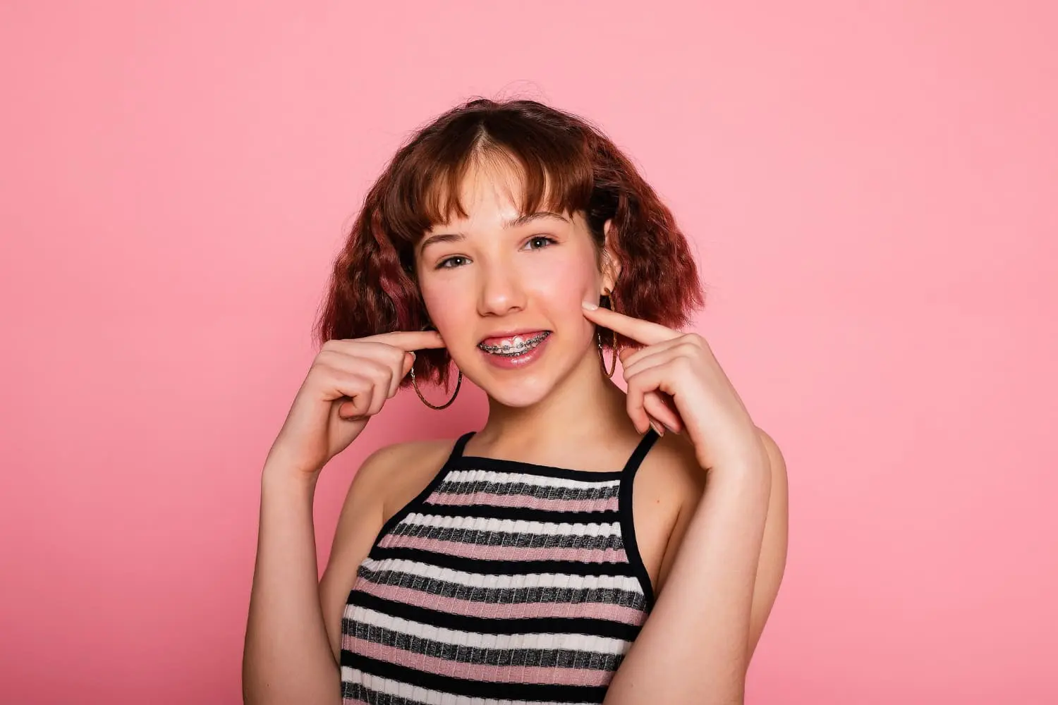 A teen girl with short brown hair, wearing braces and a striped tank top, smiles and points to her cheeks against a solid pink background in Jacksonville, FL, showcasing her confidence while highlighting the differences between Invisalign vs braces.
