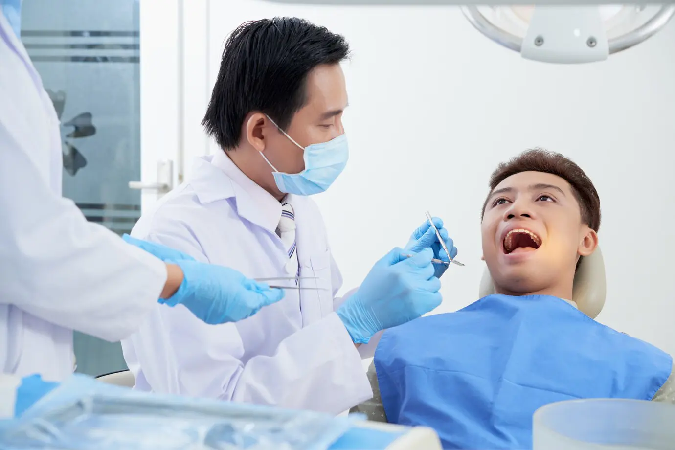 "In Jacksonville FL, a dentist wearing a mask examines a male patient’s open mouth while an assistant holds dental tools, looking for signs of tongue thrust or other oral habits that may affect alignment.