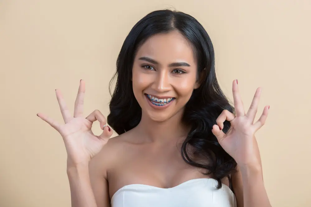 Woman with long black hair and adult braces, wearing a white strapless top, smiles and makes the "OK" hand gesture with both hands against a plain beige background in Jacksonville, FL.