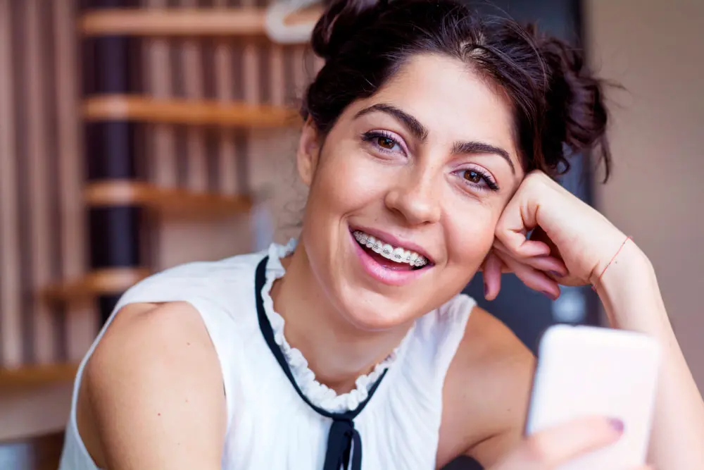 A young woman with dark hair in buns, wearing adult braces and a white sleeveless top, smiles while holding a smartphone indoors in Jacksonville, FL.