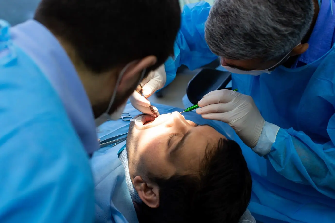 In Jacksonville FL, two dental professionals in blue gowns and gloves perform a dental procedure on a patient, carefully assessing for conditions like tongue thrust that could affect oral health.