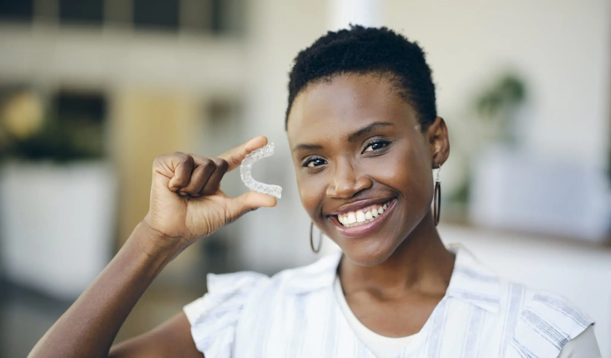 Smiling woman holding an Invisalign Clear Aligner in her right hand, standing indoors with a blurred background in Jacksonville, FL.