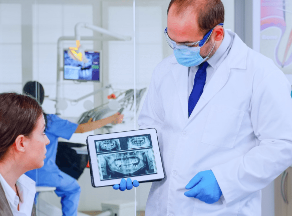 A dentist in protective gear shows a patient dental X-rays on a tablet in a clinic, while another dental professional works in the background, possibly discussing jaw surgery options in Jacksonville, FL.