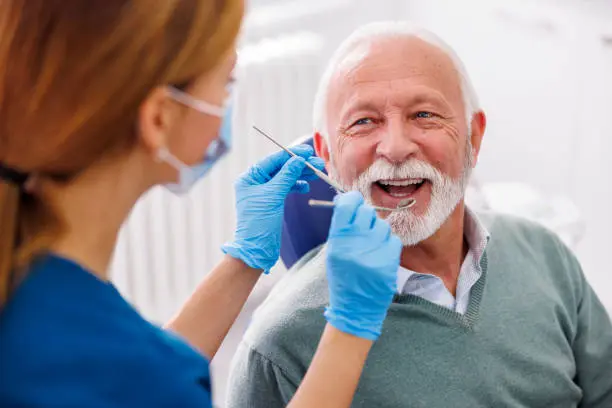 A dentist wearing gloves examines the teeth of an older man with white hair and a beard, sitting in a dental chair and smiling, possibly following up on jaw surgery in Jacksonville, FL.